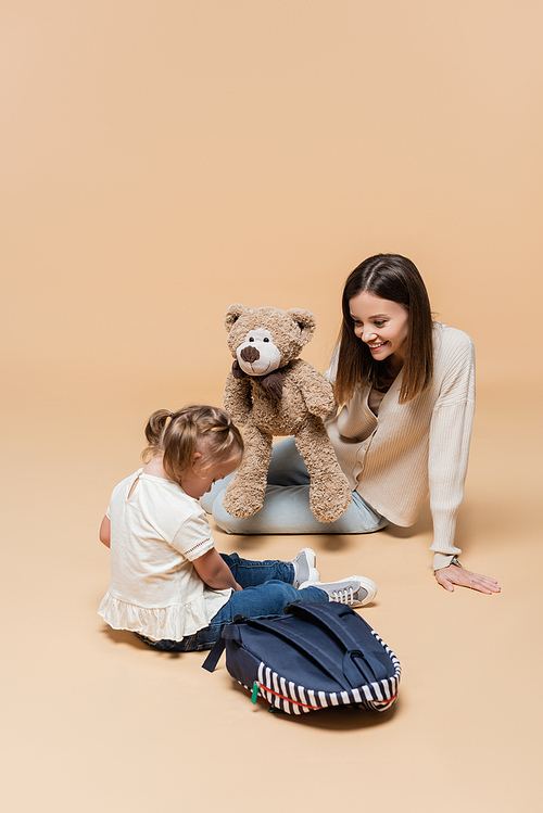 happy mother holding teddy bear near girl with down syndrome and backpack on beige