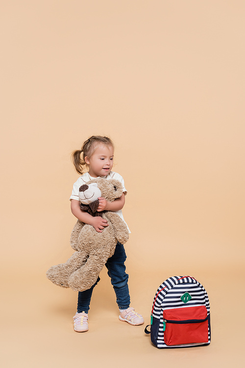 happy girl with down syndrome holding soft toy while standing near backpack on beige