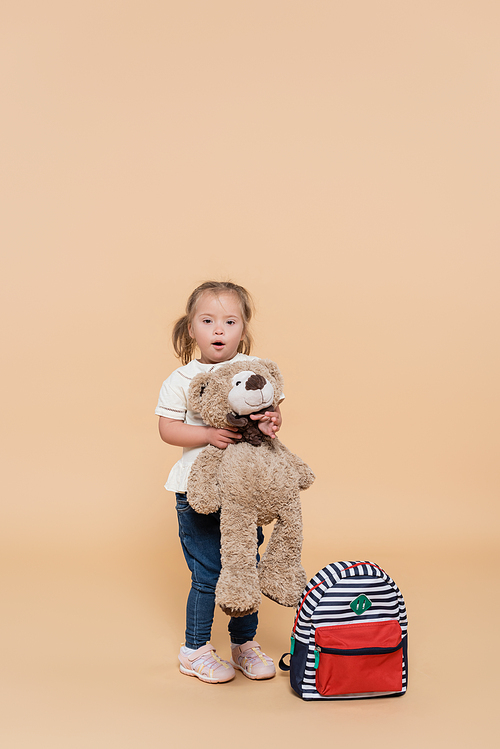 surprised girl with down syndrome holding soft toy while standing near backpack on beige