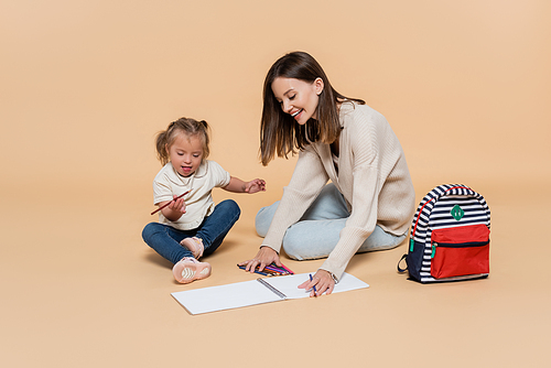 happy mother sitting near kid with down syndrome drawing near colorful pencils and backpack on beige