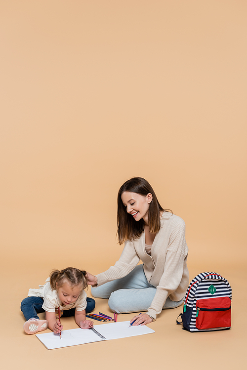 cheerful mother sitting near girl with down syndrome drawing near colorful pencils and backpack on beige