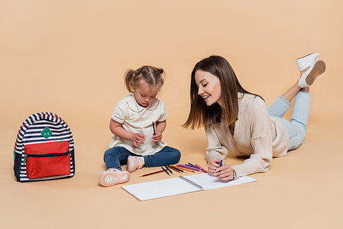joyful mother lying near kid with down syndrome drawing near colorful pencils and backpack on beige