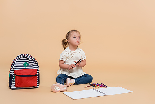 kid with down syndrome sitting near colorful pencils and backpack on beige