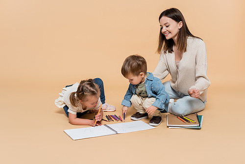 happy woman sitting near toddler boy and girl with down syndrome drawing on beige