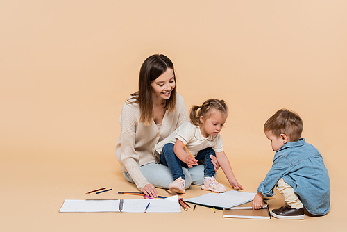 happy mother sitting near girl with down syndrome, toddler boy and colorful pencils on beige