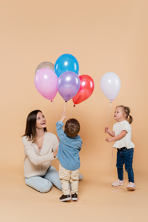 happy mother sitting near girl with down syndrome and toddler boy holding colorful balloons on beige