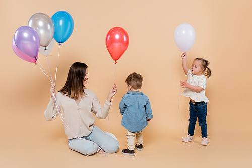 cheerful mother sitting near girl with down syndrome and toddler boy holding colorful balloons on beige
