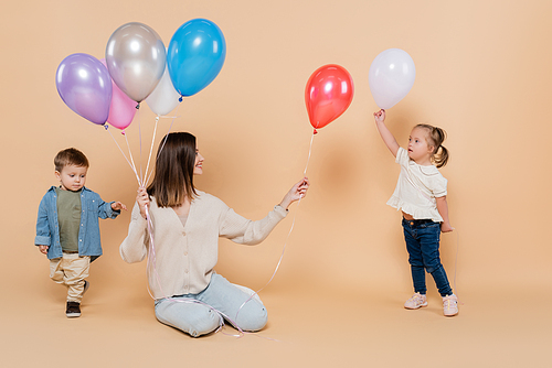 positive woman sitting near girl with down syndrome and toddler boy while holding colorful balloons on beige