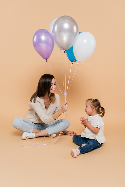 positive woman sitting near girl with down syndrome while holding colorful balloons on beige