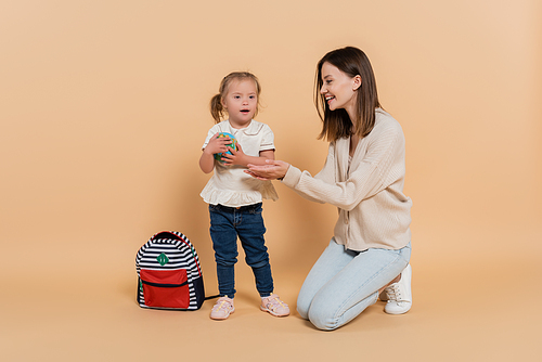 girl with down syndrome holding small globe near happy mother and backpack on beige