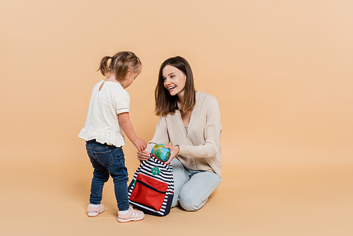 happy woman holding small globe and backpack while looking at kid with down syndrome on beige