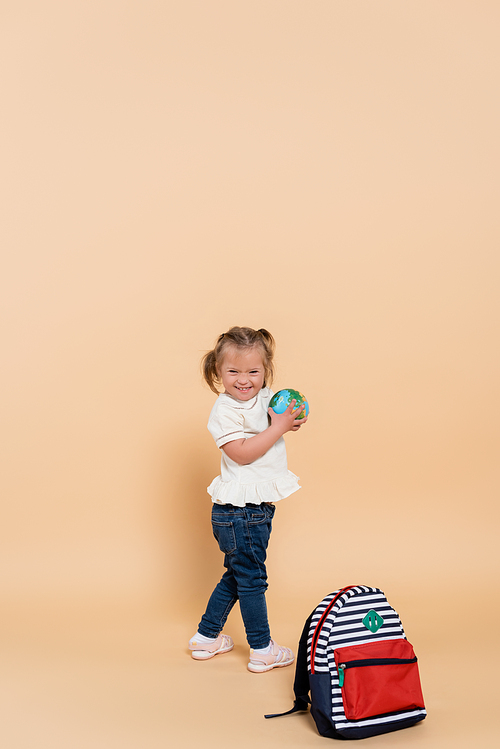 happy kid with down syndrome holding small globe near backpack on beige