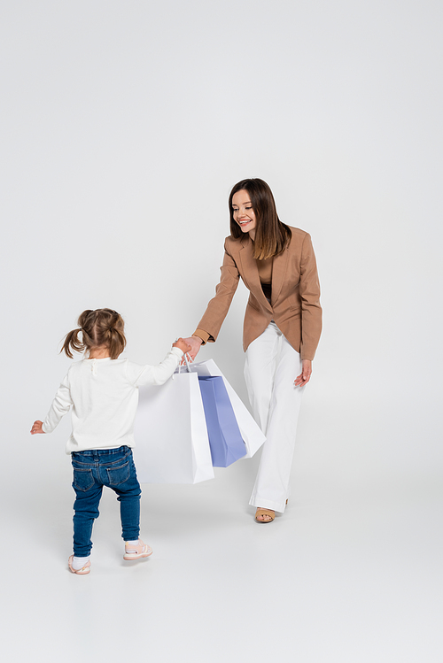 happy and stylish mother holding shopping bags while looking at kid with down syndrome on grey