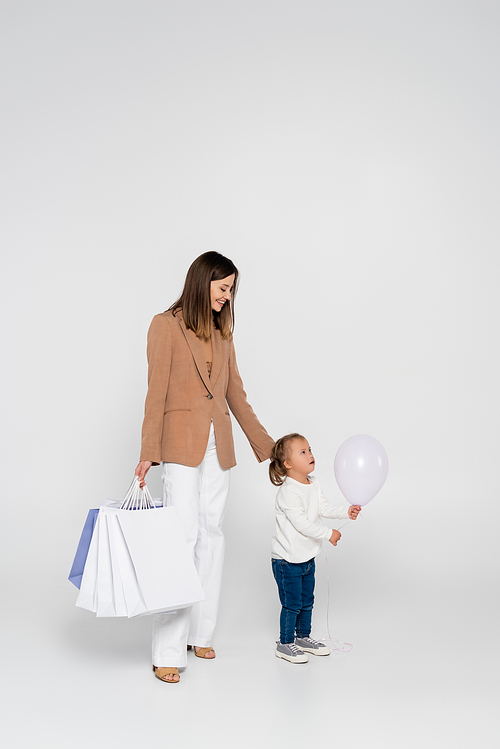 happy mother holding shopping bags and looking at girl with down syndrome near balloon on grey