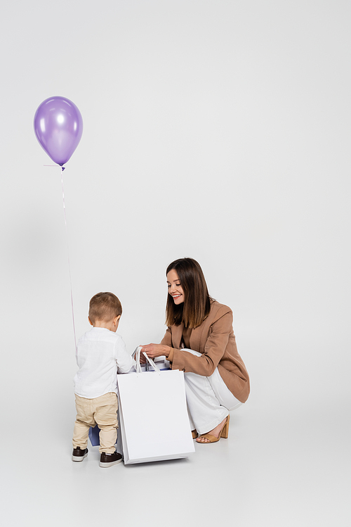 happy mother holding shopping bags near toddler son and balloon on grey