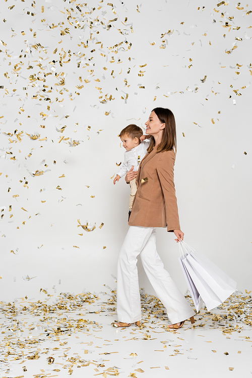 full length of happy mother holding shopping bags and toddler son in arms near confetti on grey