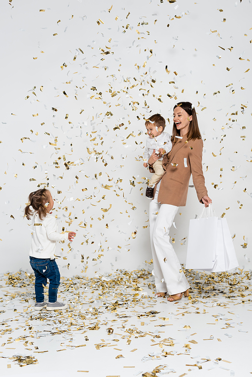 happy mother holding shopping bags and toddler boy in arms near girl with down syndrome and confetti on grey