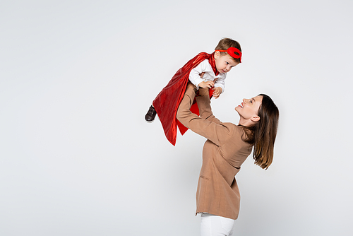 happy mother lifting toddler son in superhero costume isolated on grey