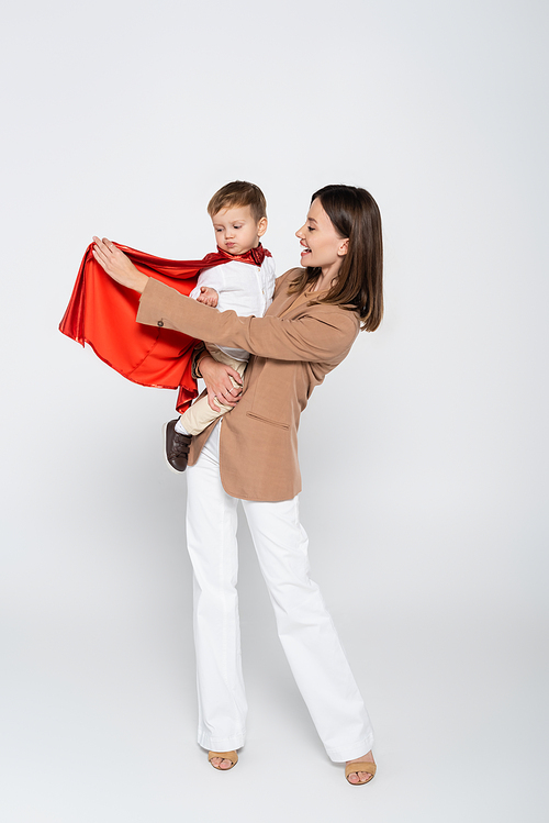 excited mother holding in arms toddler son in superhero costume on grey