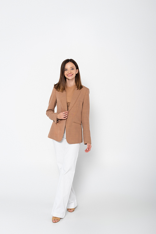 full length of happy young woman in beige blazer posing on white