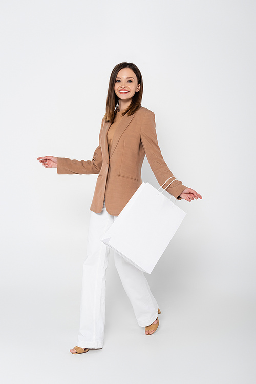 full length of joyful young woman in beige blazer holding shopping bag while posing on white