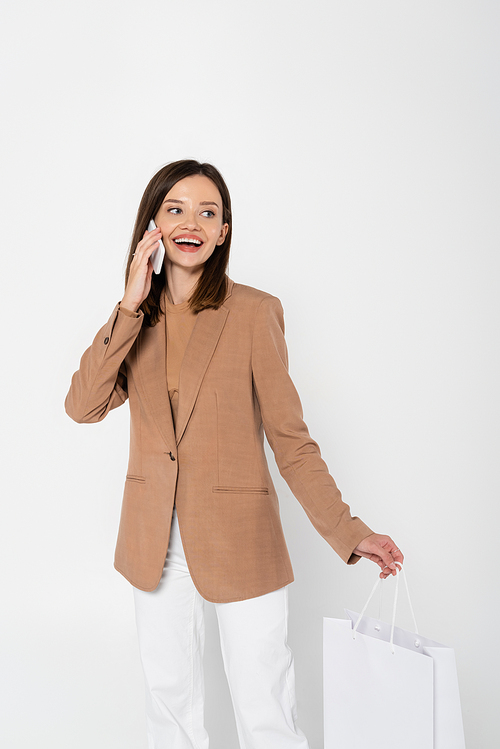 happy young woman in beige blazer holding shopping bag and taking on smartphone on grey