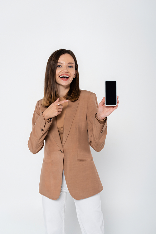 happy young woman in beige blazer pointing at smartphone with blank screen on grey