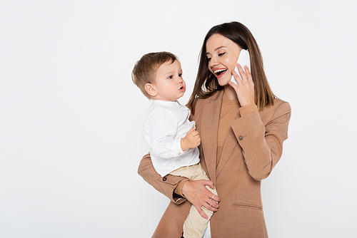 happy young woman in beige blazer talking on smartphone and holding in arms toddler boy isolated on grey