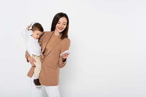 happy woman in beige blazer holding smartphone and toddler boy in headphones isolated on grey