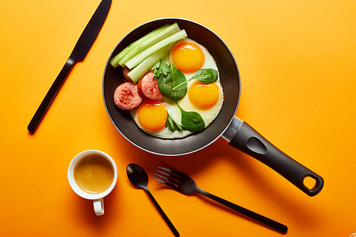 top view of fried eggs with spinach leaves, cucumber and sausage in frying pan near coffee, cutlery on orange background