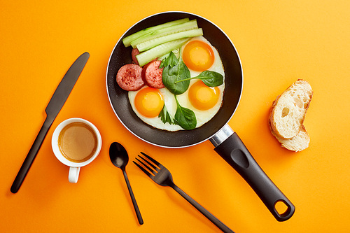 top view of fried eggs with spinach leaves, cucumber and sausage in frying pan near coffee, cutlery and bread on orange background