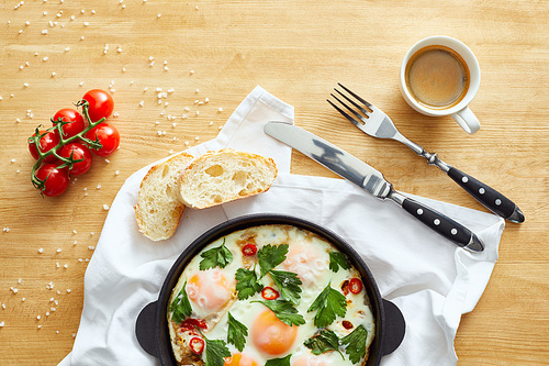 top view of fried eggs in pan near bread, cutlery, coffee and tomatoes on napkin on wooden table
