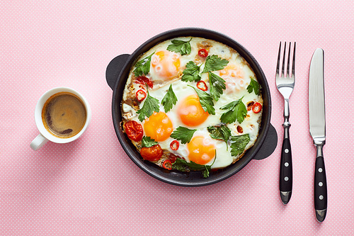 top view of fried eggs with parsley, tomatoes and chili pepper in frying pan near cutlery and coffee on pink background