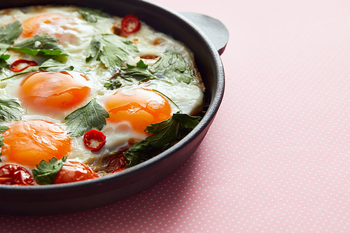 close up view of fried eggs with parsley, tomatoes and chili pepper in frying pan on pink background
