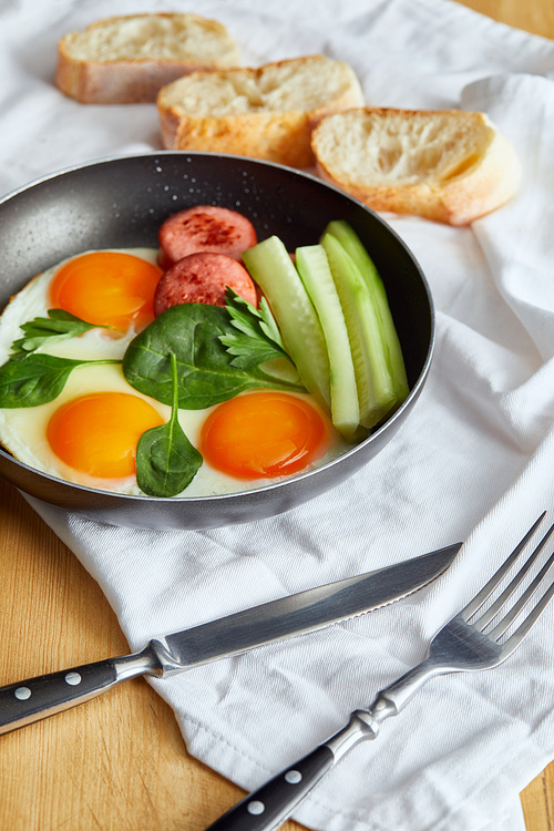 selective focus of fried eggs in frying pan with spinach, cucumber and sausage at wooden table with cutlery and bread on napkin