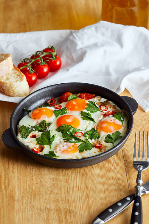 fried eggs with parsley and chili pepper in pan near napkin, bread, tomatoes and napkin on wooden table