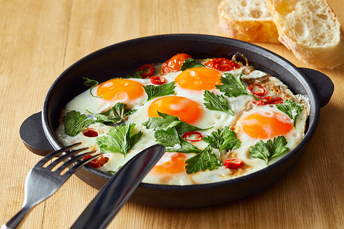 fried eggs with parsley and chili pepper on wooden table with cutlery and bread