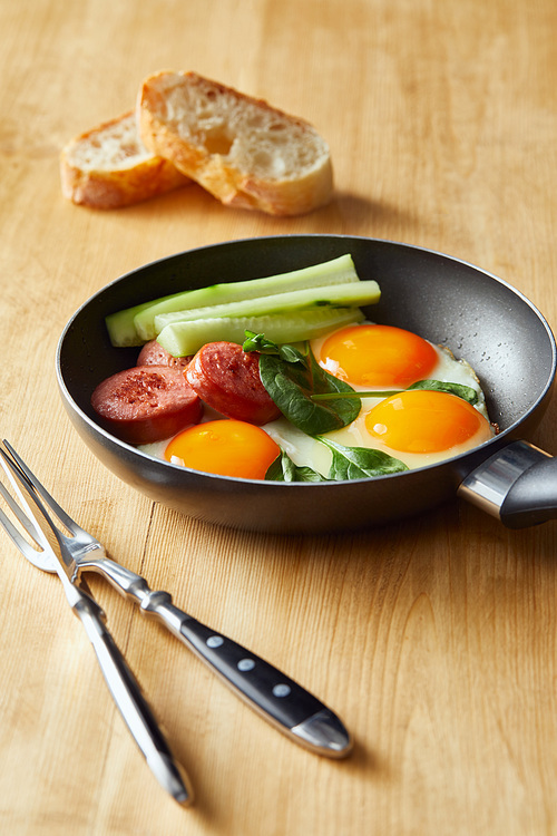 selective focus of fried eggs in frying pan with spinach, cucumber and sausage at wooden table with cutlery and bread