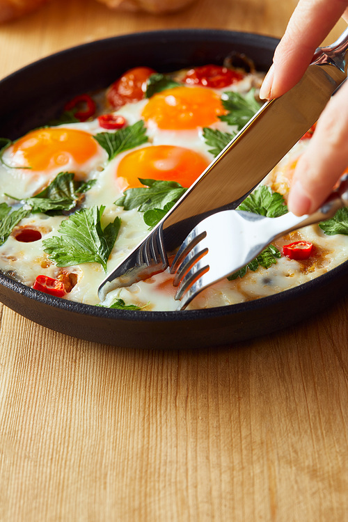 cropped view of eating fried eggs with parsley and chili pepper on wooden table with fork and knife