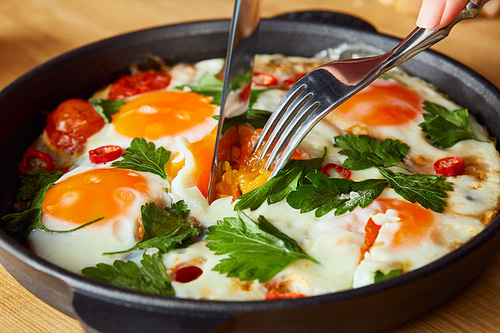 cropped view of eating fried eggs with parsley and chili pepper on wooden table with fork and knife