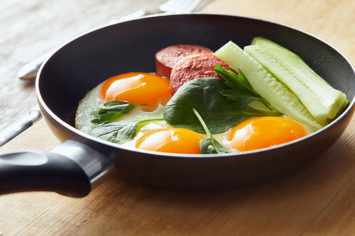 fried eggs in frying pan with spinach, cucumber and sausage at wooden table