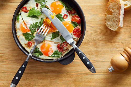 top view of fried eggs with parsley and chili pepper on wooden table with cutlery and bread
