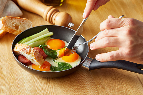 cropped view of man eating fried eggs with fork and knife at wooden table