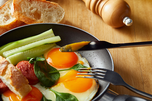 fried eggs in frying pan with vegetables and sausage near fork, knife, bread at wooden table