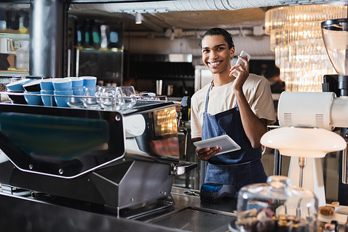 Smiling african american barista holding devices near coffee machine in cafe
