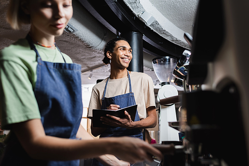 Smiling african american barista in apron writing on notebook near blurred colleague and coffee machine in cafe