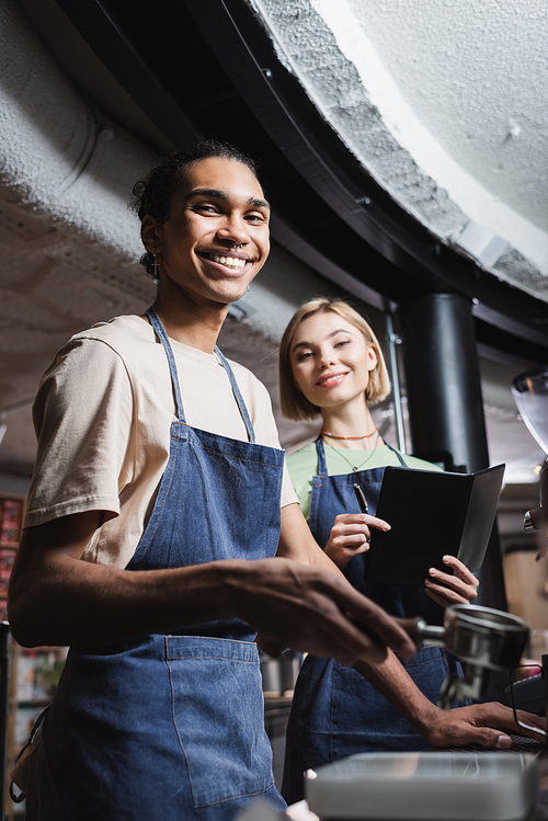 Smiling multiethnic baristas in aprons holding notebook and coffee portafilter in cafe