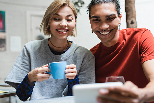 Smiling interracial couple with coffee looking at blurred smartphone in cafe