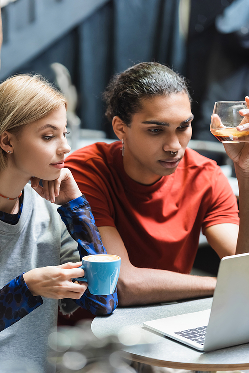 Young interracial freelancers holding coffee near laptop in cafe