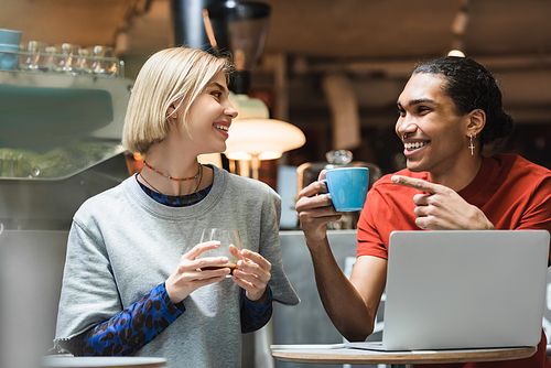Positive african american freelancer holding coffee and pointing at friend near devices in cafe